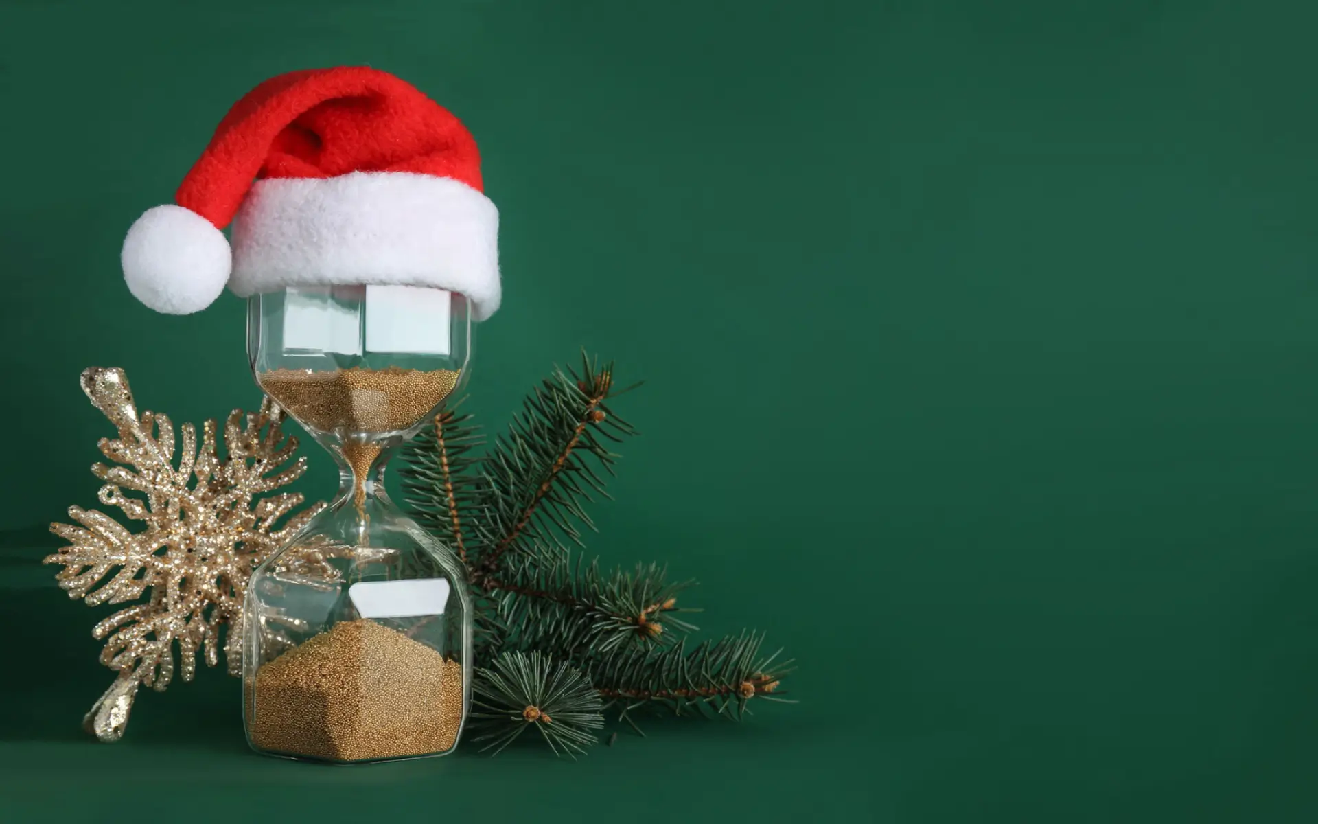 Sanduhr auf grünem Hintergrund. Die Sanduhr hat eine Weihnachtsmütze auf. Hinter der Sanduhr liegt Weihnachtsbaumschmuck.